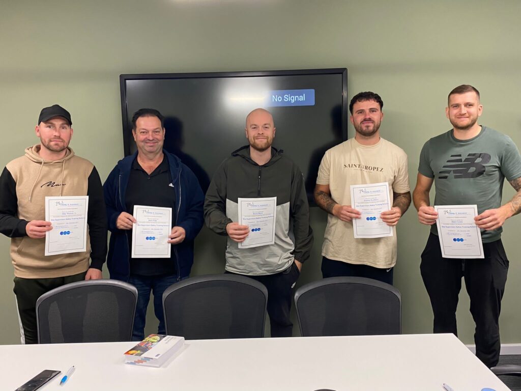 Five men stand in a row, facing the camera and holding their SMSTS course certificates. They are in a conference room and are all smiling.